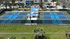 A tennis court with multiple courts, surrounded by a fence, and located near a beach with buildings in the background.