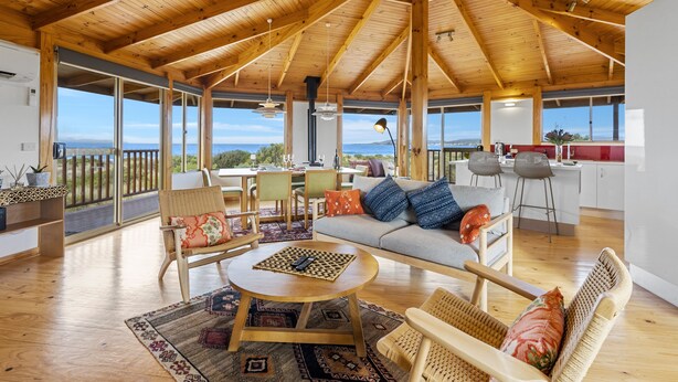 A spacious living area with a high wooden ceiling, large windows offering a view of the outdoors, a cozy seating area with a sofa and chairs, and a kitchen in the background. - Peace & Plenty - beachfront hideaway with an indoor heated pool (Dolphin Sands)