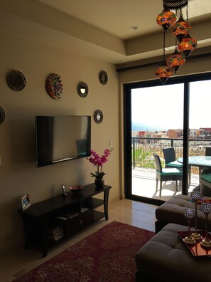 Una sala de estar con un televisor de pantalla plana, placas decorativas en la pared, un jarrón con flores, un tapete rojo estampado y un balcón con mobiliario exterior.