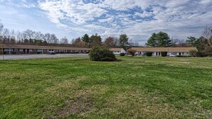 A motel with a grassy lawn, a parking lot, and a row of trees in the background. - Newport Motel (Newport)