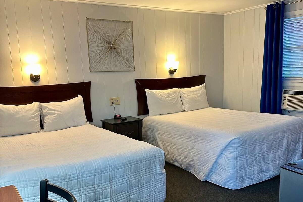 Een hotelkamer met twee bedden, een bureau, een stoel, een klein tafeltje, een nachtkastje, een wandklok en een raam met gordijnen.