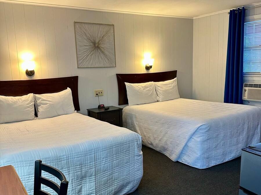 Habitación de hotel con dos camas, un escritorio, una silla, una mesita, una mesita de noche, un reloj de pared y un ventanal con cortinas.