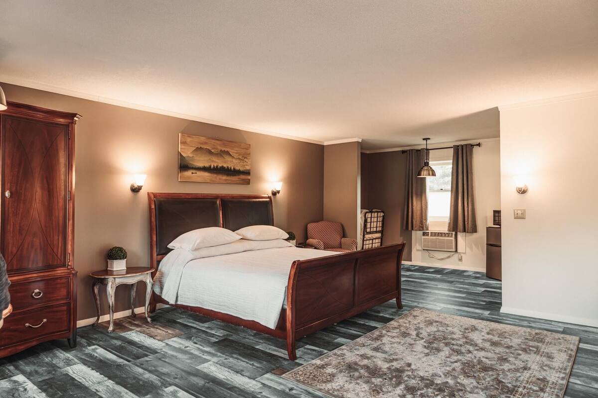 Een slaapkamer met een houten bed, een klein bijzettafeltje, een nachtkastje, een lamp, een schilderij aan de muur en een raam met gordijnen.