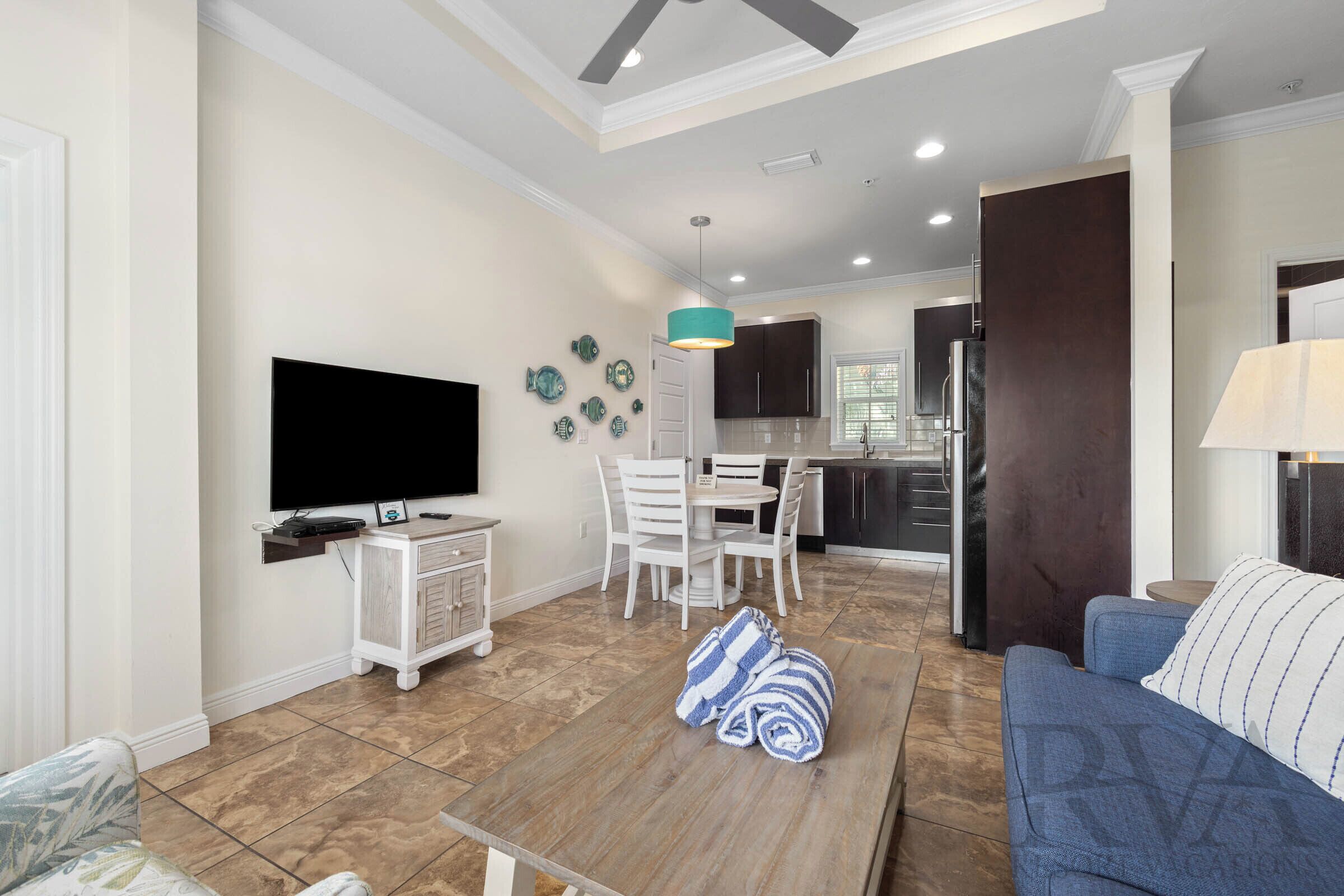 A modern living room with a flat-screen TV, a dining area with a table and chairs, a kitchen in the background, and a blue sofa.