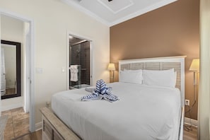 A hotel room with a large bed, two bedside lamps, a bathroom with a glass door, and a wooden bench.