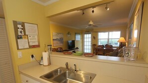 A kitchen with a stainless steel sink, a bulletin board with notices, a television, and a dining area with chairs and a table. - Ground-Floor Gulf Retreat-Outrigger #5 by RVA (Longboat Key)