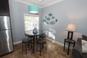 A kitchen with a stainless steel refrigerator, a round dining table with four stools, a teal pendant light, and a wall with decorative fish plates. - Relax & Unwind – 1BR Beach Club Siesta Key 110D RVA (Sarasota)