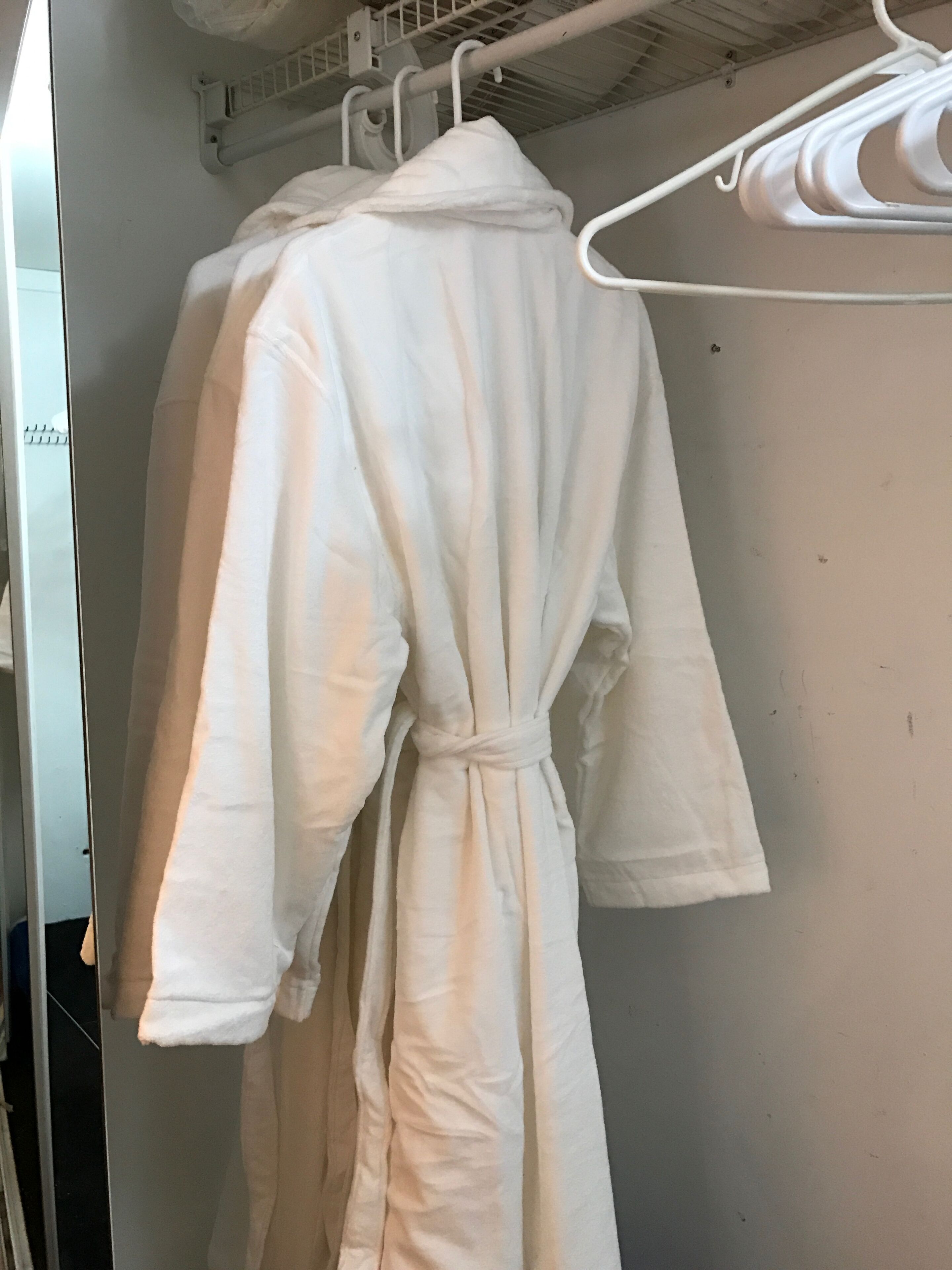 A white robe hanging on a white metal hanger in a room with a white wall and a ceiling with visible pipes.