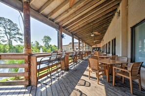 Un patio al aire libre con mesas y sillas de madera, barandilla de madera y vista al agua y los árboles.