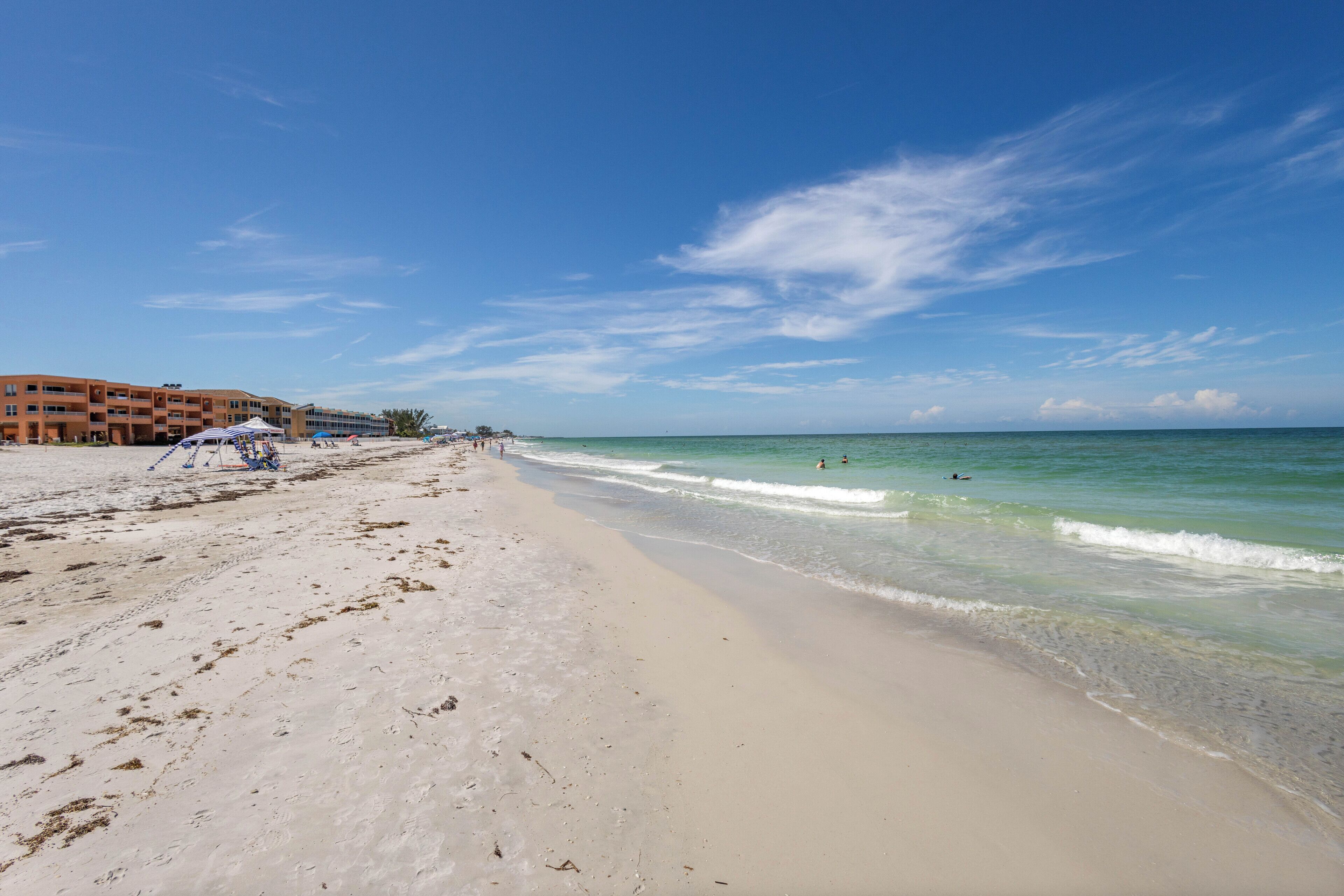 A beach with a hotel in the background, clear skies, and calm sea.
