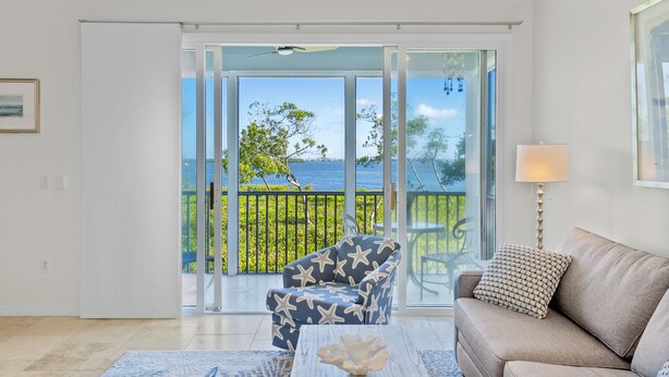 A living room with a sofa, armchair, and coffee table, a balcony with a view of the sea, and a lamp. - Play, Stay & Bring Your Pup: Cedars East 832 RVA (Longboat Key)