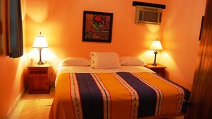 Een slaapkamer met een bed, twee bedlampen, een kunstwerk in een lijst aan de muur, en een airconditioning boven het bed.
