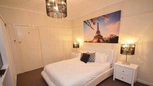 Een slaapkamer met een wit bed, twee nachtkastjes met lampen, een schilderij van de Eiffeltoren en een deur.