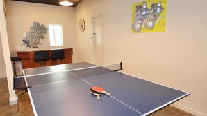Een tafeltennistafel in een ruimte met een houten kast, stoelen, een raam en een gele afbeelding aan de muur.
