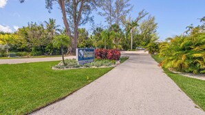 A pathway leading to a sign for Outrigger Resort, surrounded by lush greenery and palm trees. - 2nd-Floor Gulffront Outrigger #6 Condo by RVA (Longboat Key)