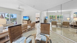 A spacious living area with wicker furniture, a television, and a glass wall offering a view of the outdoors. - Coastal Serenity Sea Twig 102-Beachfront Condo w Private Lanai & Pool by RVA (Longboat Key)