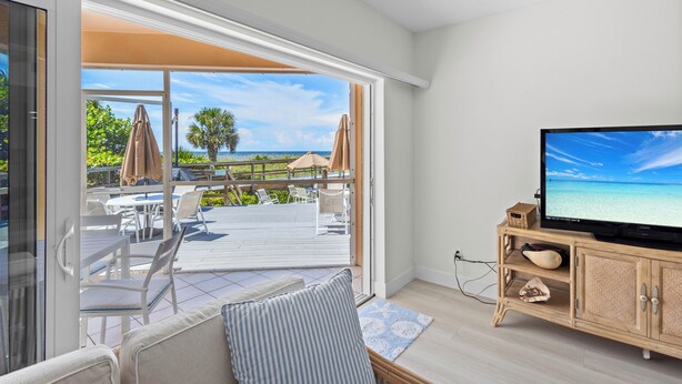 A room with a TV, a sofa, and a sliding glass door leading to an outdoor deck with chairs and umbrellas. - Coastal Serenity Sea Twig 102-Beachfront Condo w Private Lanai & Pool by RVA (Longboat Key)