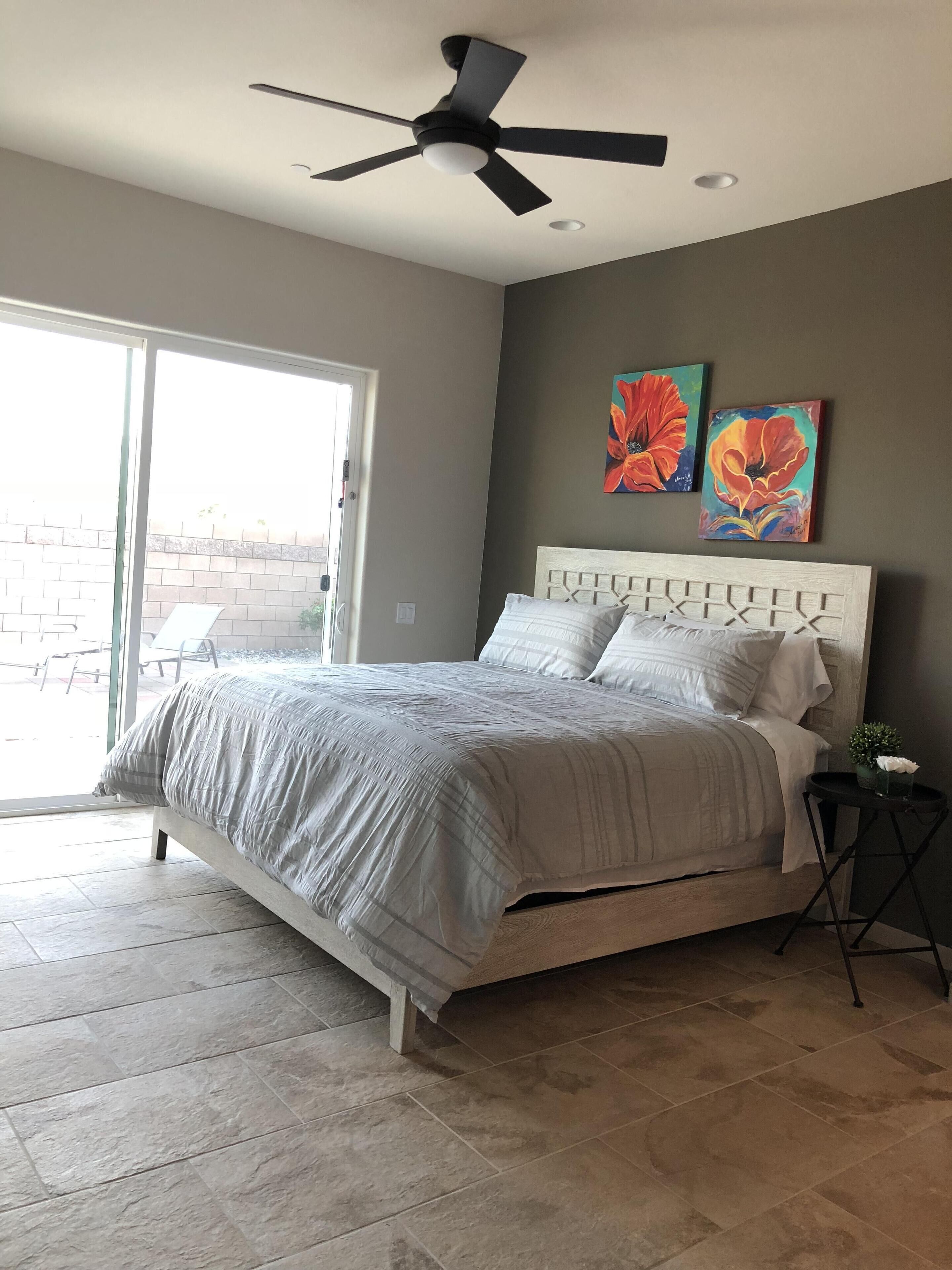 A bedroom with a bed, a ceiling fan, a window, and two paintings on the wall.