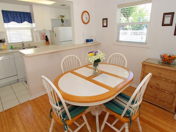 A compact kitchen with white cabinets, a wooden dining table with chairs, a clock, and a window with blinds. - Siesta Key Getaway – Pet Friendly & Steps to Beach: Siesta Bungalow by RVA (Sarasota)