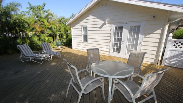 A wooden deck with a round table and four chairs, surrounded by a white house with a garage door and a small porch. - Siesta Key Getaway – Pet Friendly & Steps to Beach: Siesta Bungalow by RVA (Sarasota)