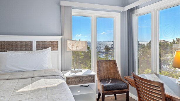 A hotel room with a bed, a chair, a desk, and large windows offering a view of the sea.