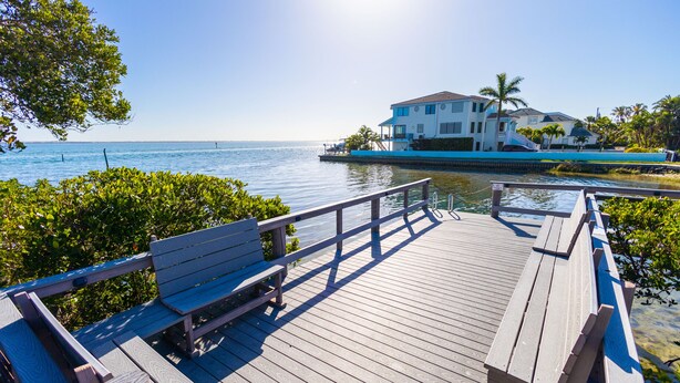 A wooden deck with benches overlooking a body of water, a white building with a balcony, and palm trees in the background. - Courtside Getaway – Cedars East #605 by RVA (Longboat Key)