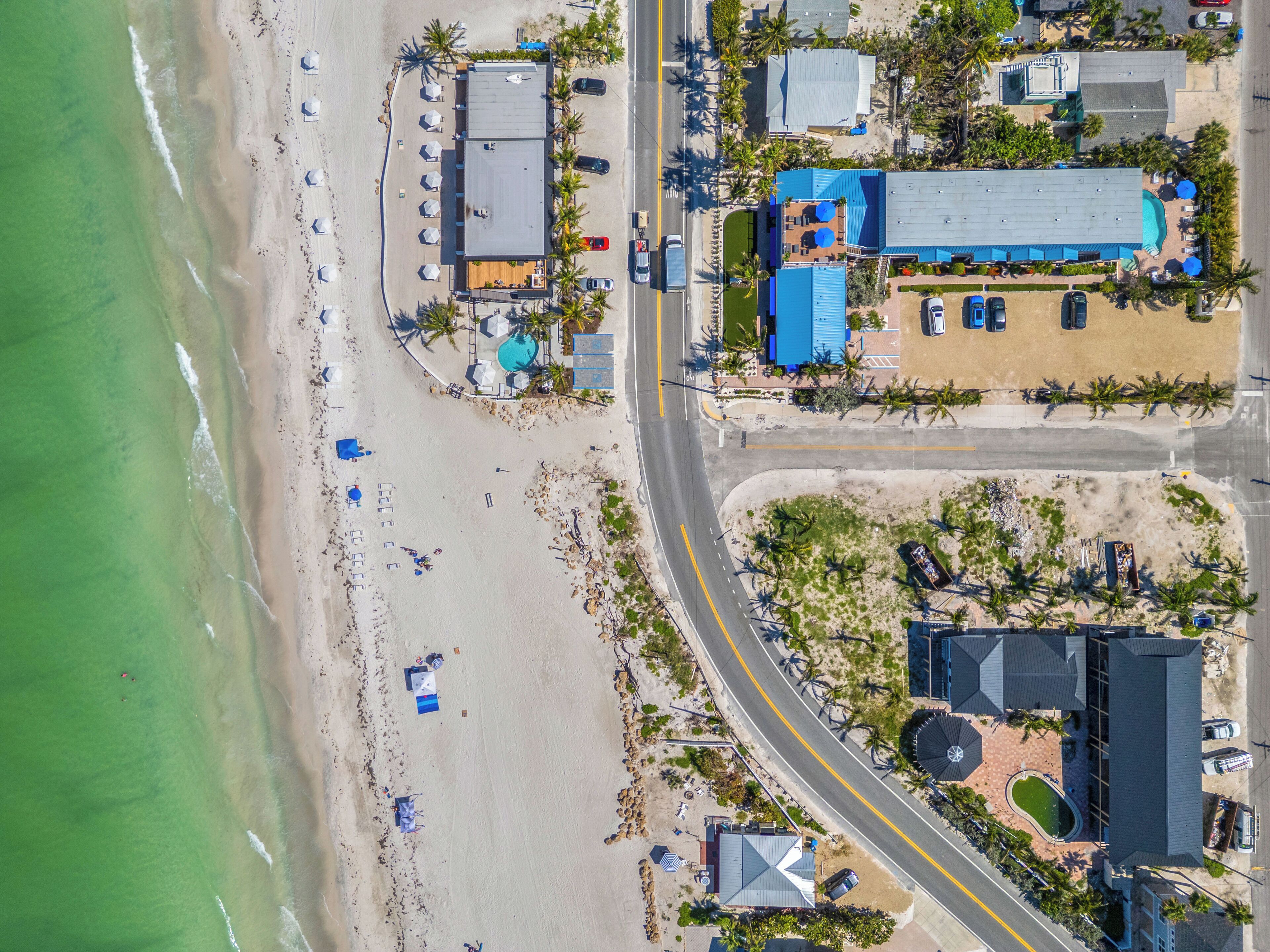 An aerial view of a coastal hotel with a swimming pool, beach umbrellas, and a parking area.