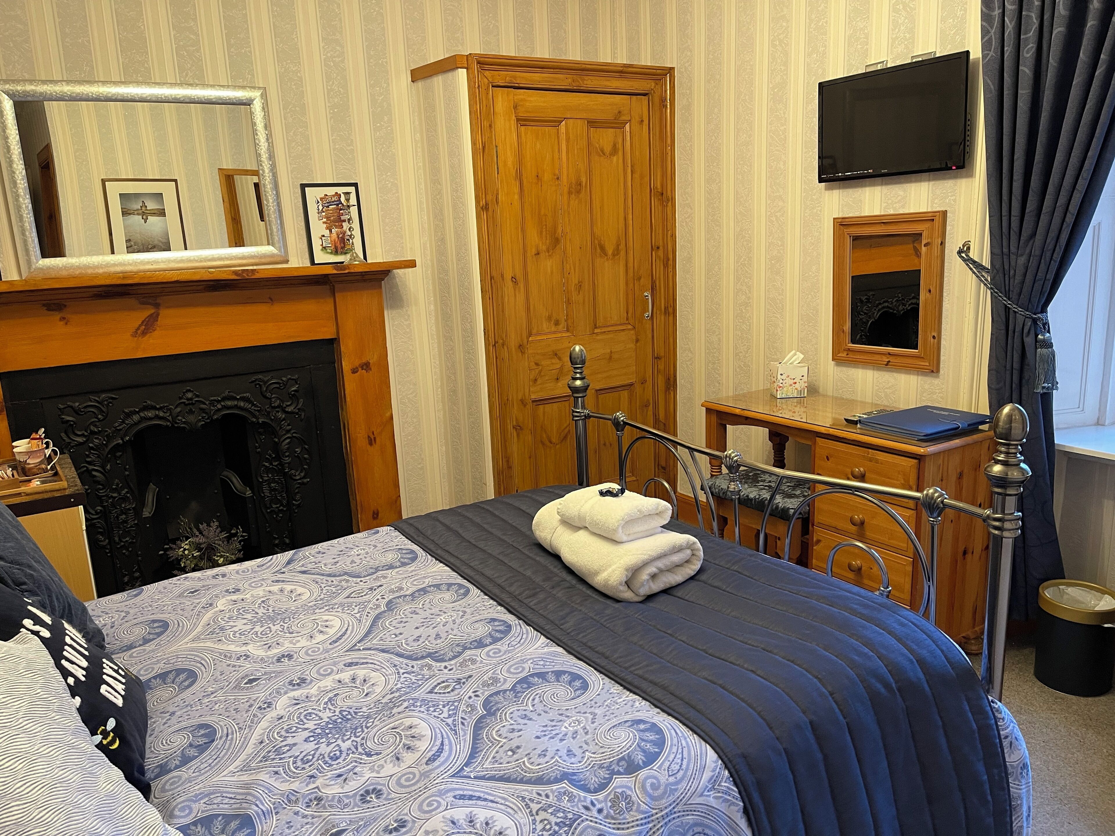 A bedroom with a bed, a wooden wardrobe, a television, a fireplace, and a desk with a laptop.
