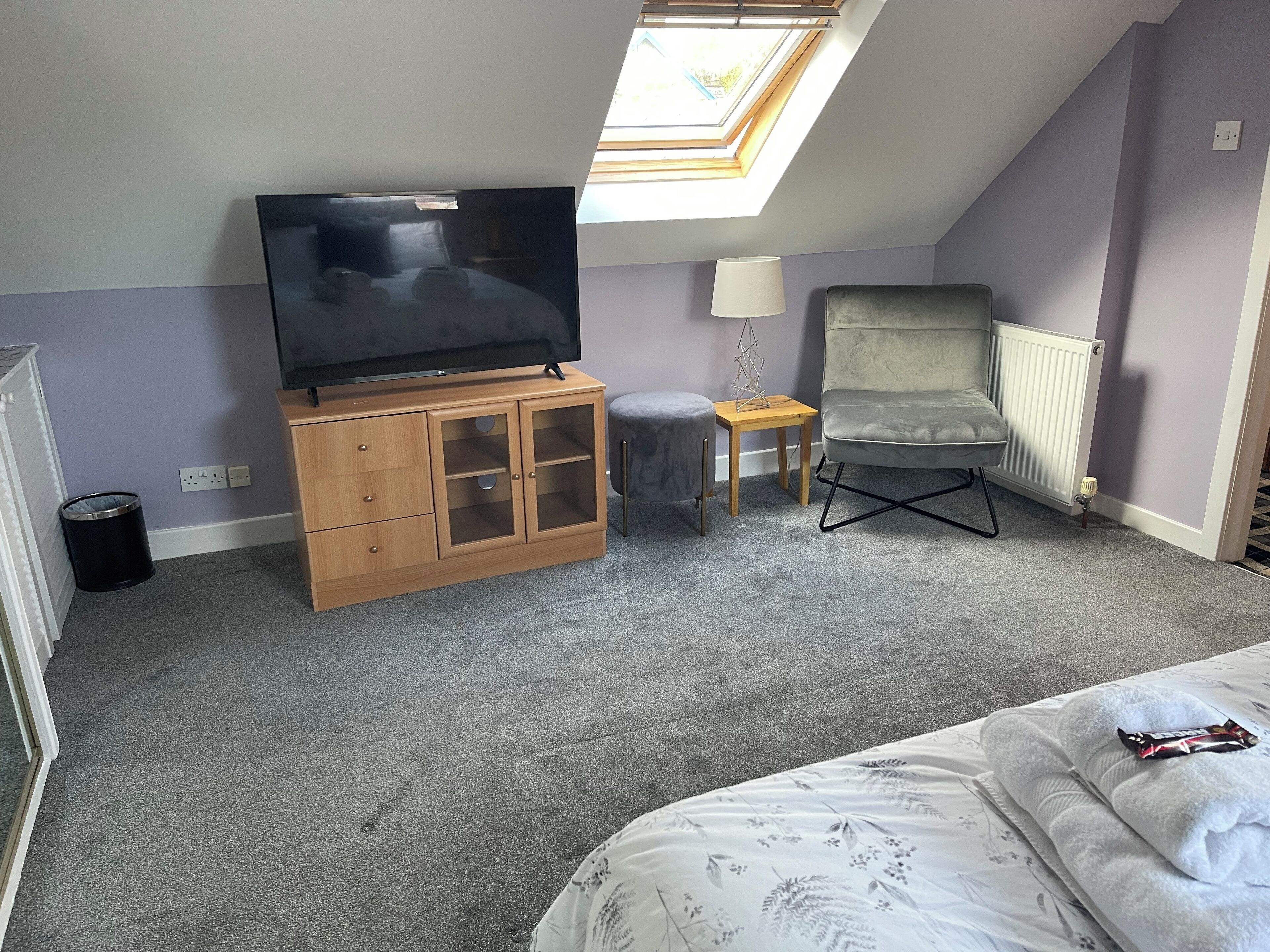 A bedroom with a bed, a TV on a wooden cabinet, a grey armchair, a small table with a lamp, and a radiator.