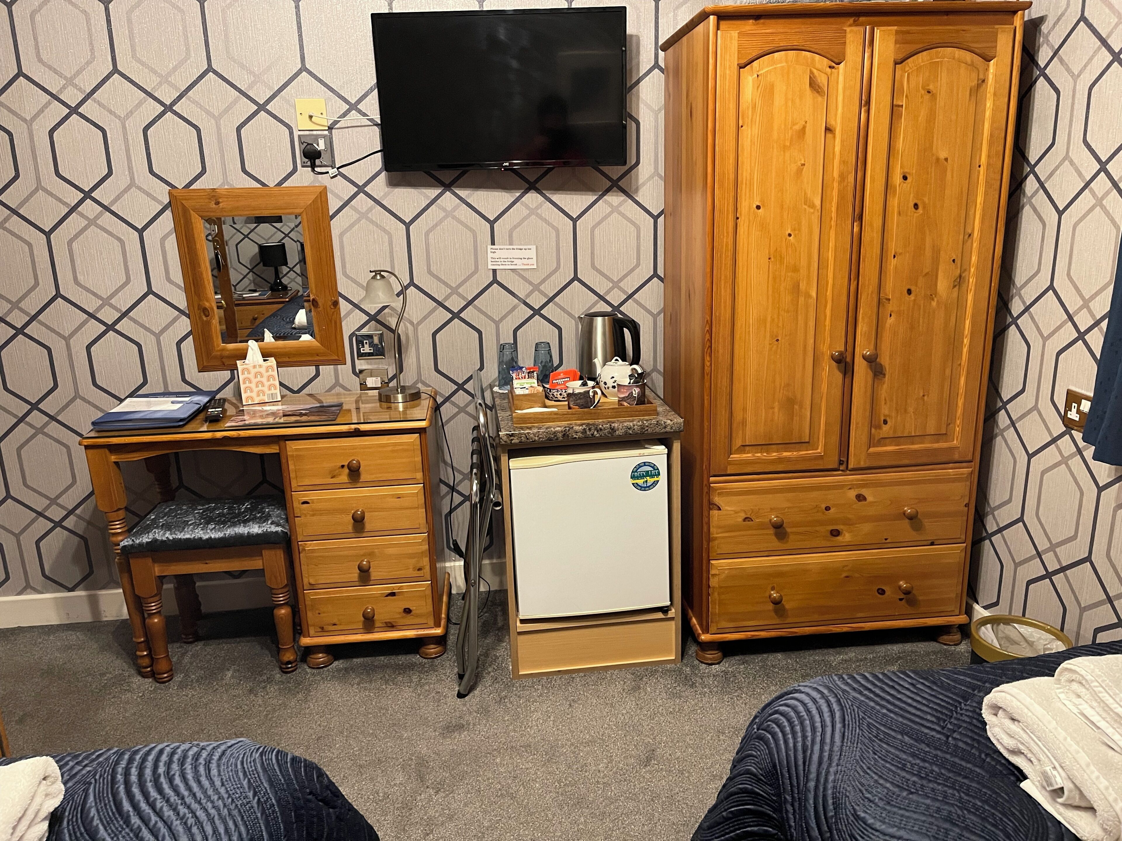 A bedroom with a wooden desk, a small refrigerator, a wardrobe, a television, and a patterned wall.