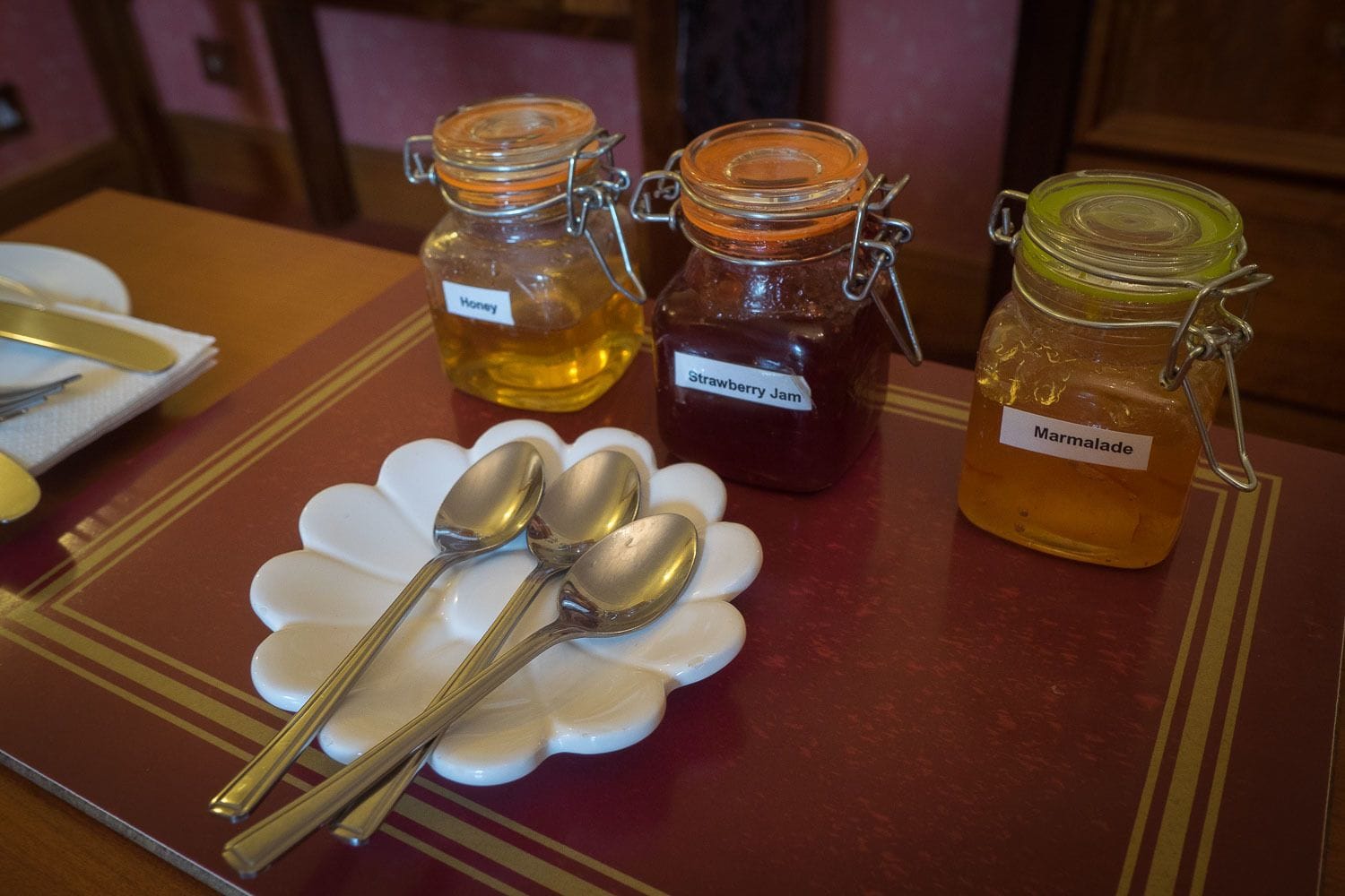 Pöydällä on kolme lasipurkkia, joissa on eri lajeja hilloja tai marmeladeja. Purkkien päällä on valkoinen, kukkakuviollinen alusta ja vieressä kaksi lusikkaa.