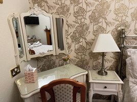 Un baño con espejo, tocador de vidrio, silla, mesita de noche con lámpara y papel tapiz con motivos florales.