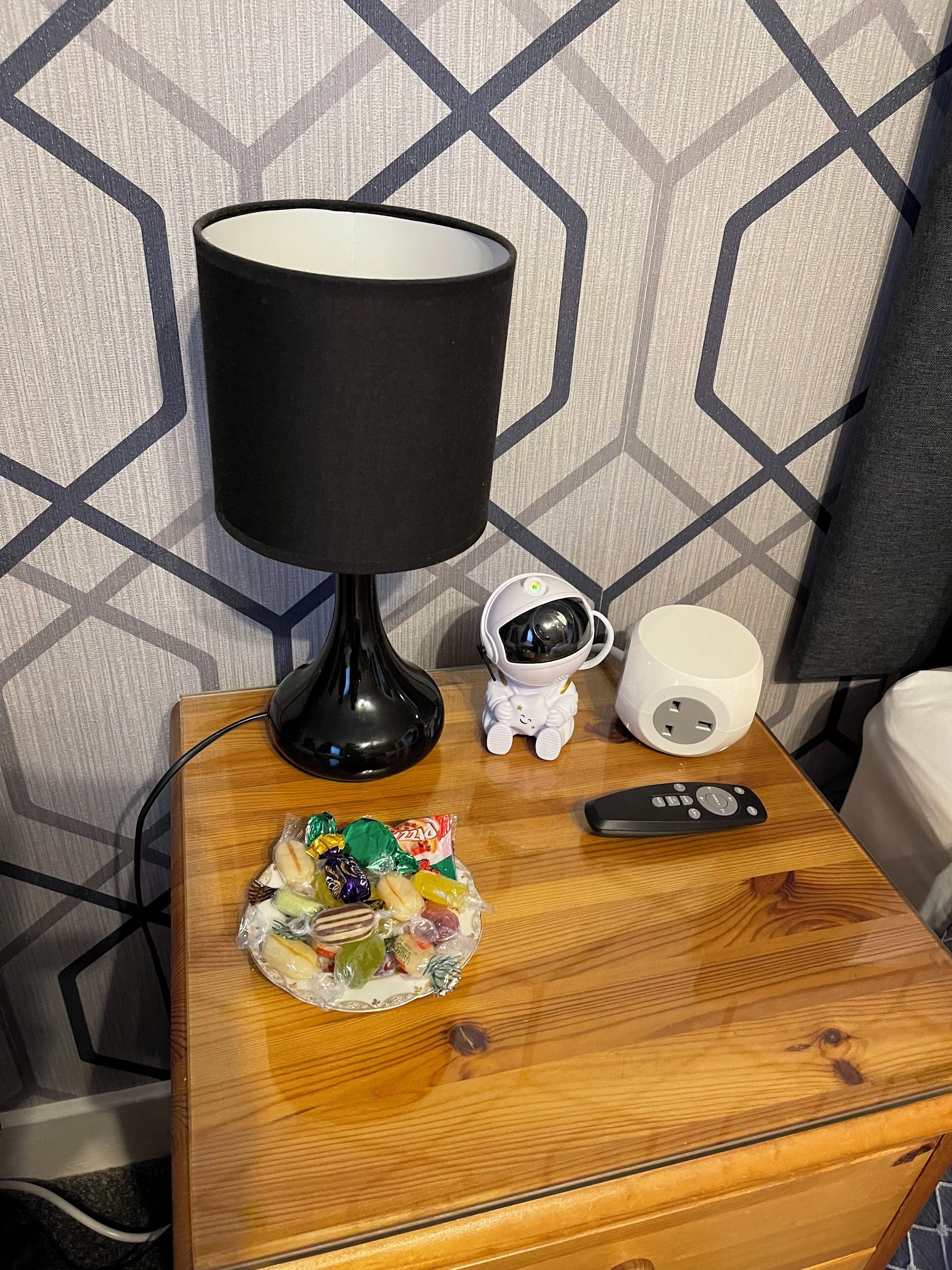 A wooden bedside table with a black lamp, a white cat-shaped night light, a white cup, a remote control, and a bowl of assorted candies.