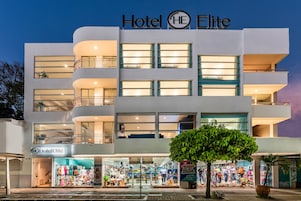 Un hotel de varios pisos con balcones y ventanas amplias, una tienda con maniquíes y un letrero que dice 'Hotel Elite'.