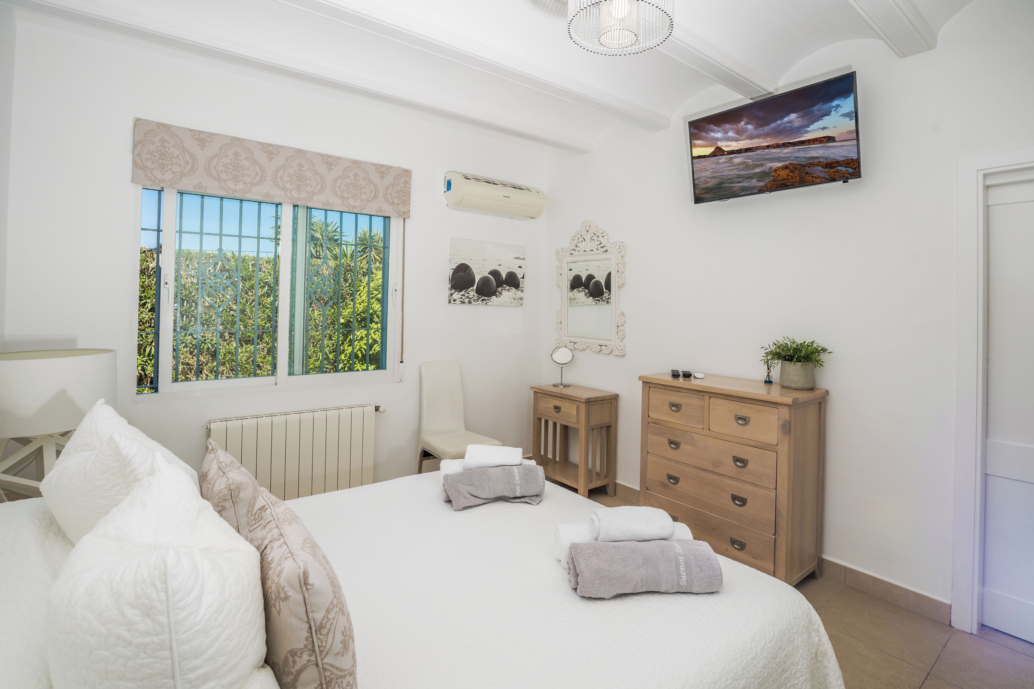 Un dormitorio con cama, cómoda, silla, espejo y televisor en la pared.