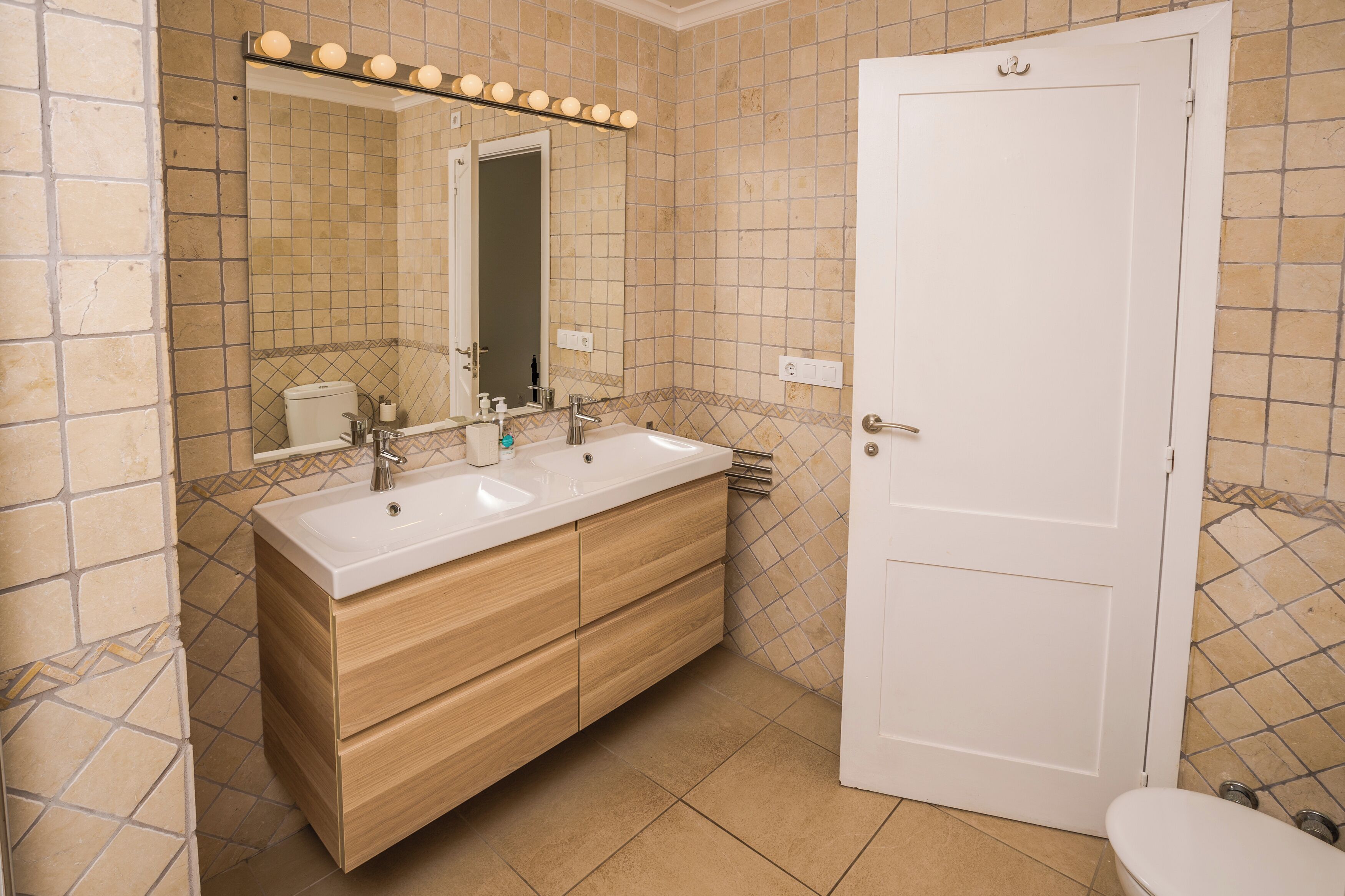 Un baño con tocador de doble lavabo, un espejo con iluminación, inodoro y puerta.