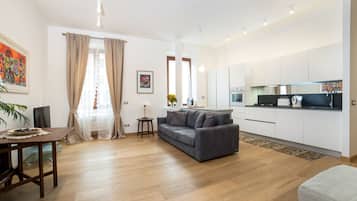 A modern living room with a sofa, a dining table, and a kitchen area with white cabinets and appliances.