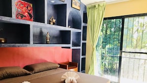 Ein Zimmer mit einem Bett, Kissen, einem Nachttisch, einem Regal mit Dekoration und Blick auf Grün durch ein Fenster.