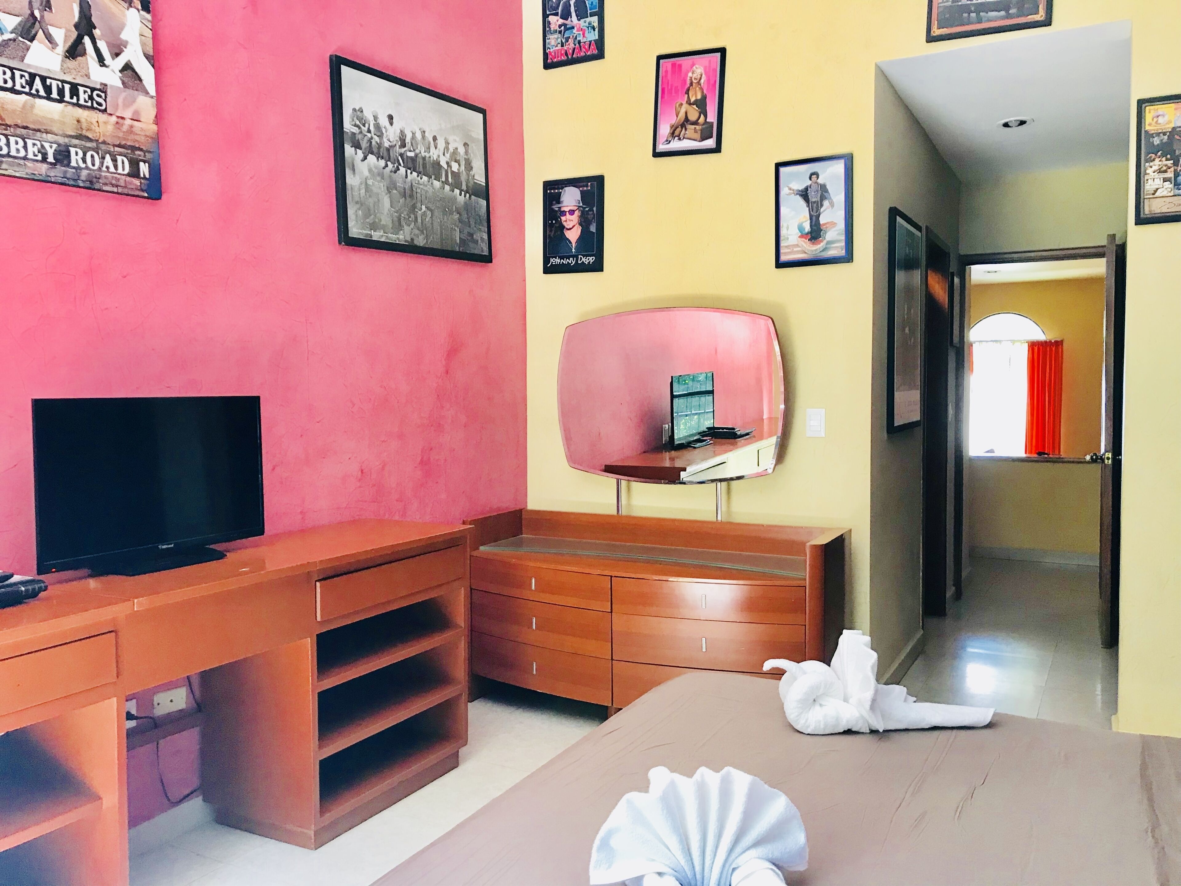 Une chambre d’hôtel dotée d’un téléviseur à écran plat, de meubles en bois, d’un mur rose et de divers tableaux encadrés.