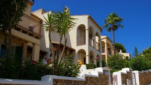 A multi-story building with a stone wall in front, balconies, and a clear blue sky. - 2 Bedroom Apartment- Ona Aucanada Alcudia, Mallorca (Aucanada, Puerto de Alcudia, Illes Balears)