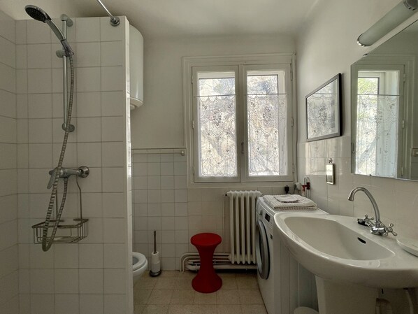 Ein Badezimmer mit Dusche, Waschbecken, Toilette, einem roten Hocker und einem Fenster mit Spitze.