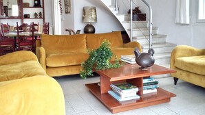 Ein Wohnzimmer mit einem gelben Sofa, einem Couchtisch mit Büchern, einer Treppe und einem Essbereich mit roten Stühlen.