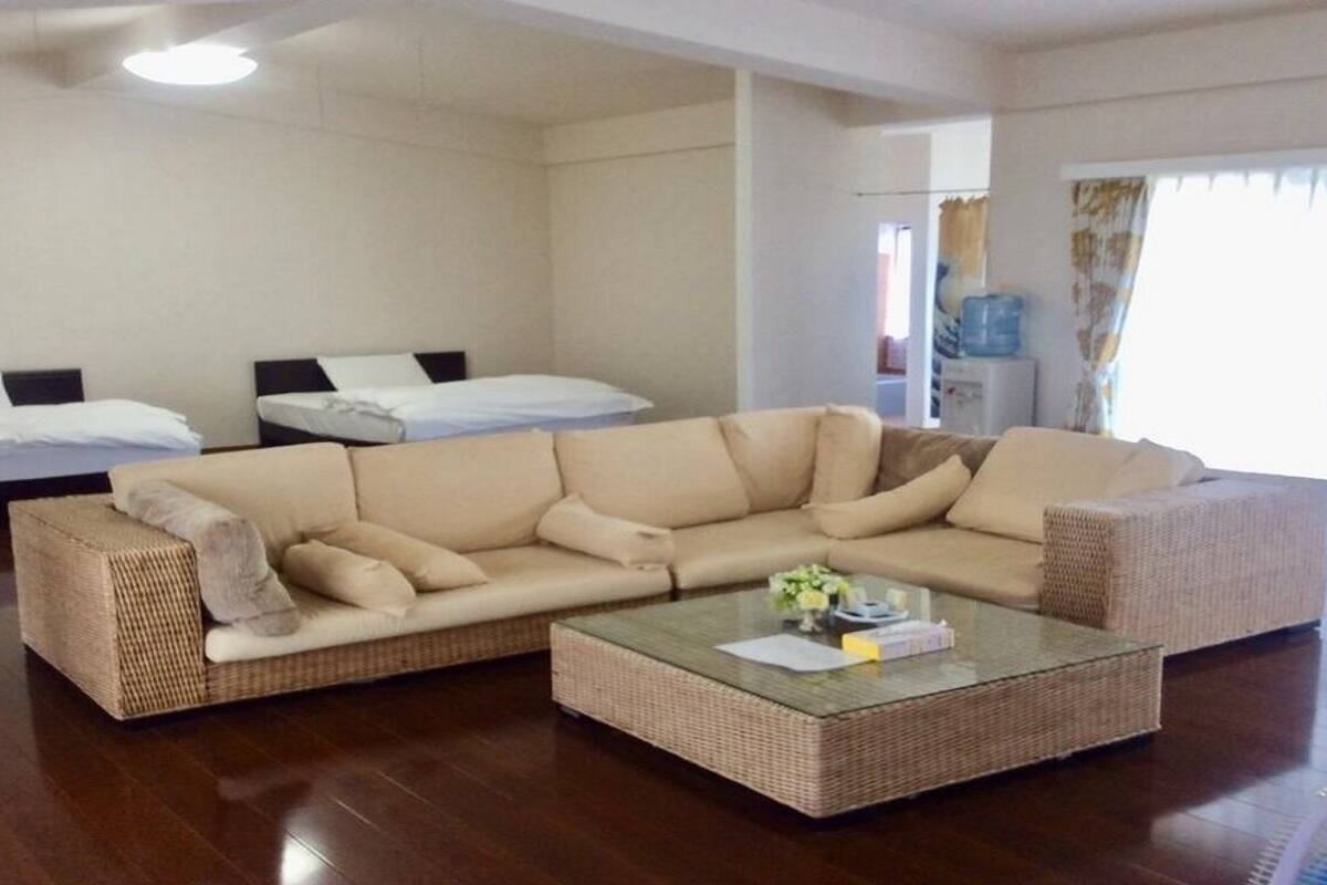 A room with a large beige sofa, a glass coffee table, a bed with white bedding, and a water dispenser.