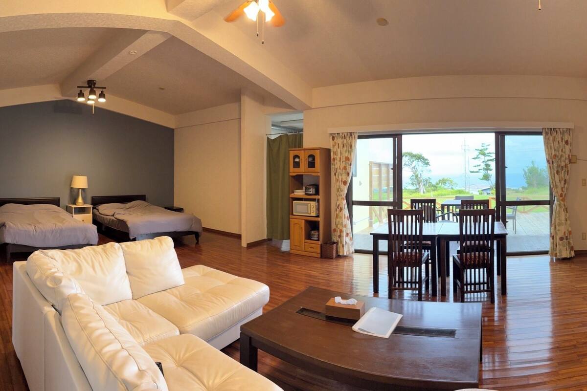 A hotel room with a sofa, a bed, a dining table with chairs, and a balcony with a view.