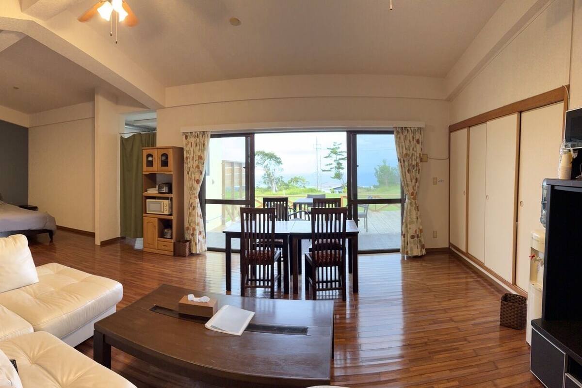 A hotel room with a sofa, two single beds, a dining area with chairs, a TV, and a view of the outdoors.