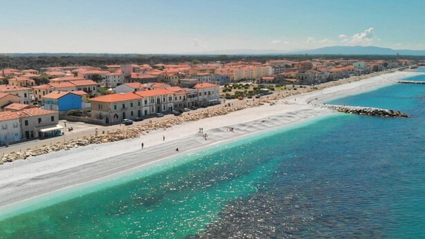 Un pueblo costero con una playa, edificios de techo rojo y un mar azul cristalino.