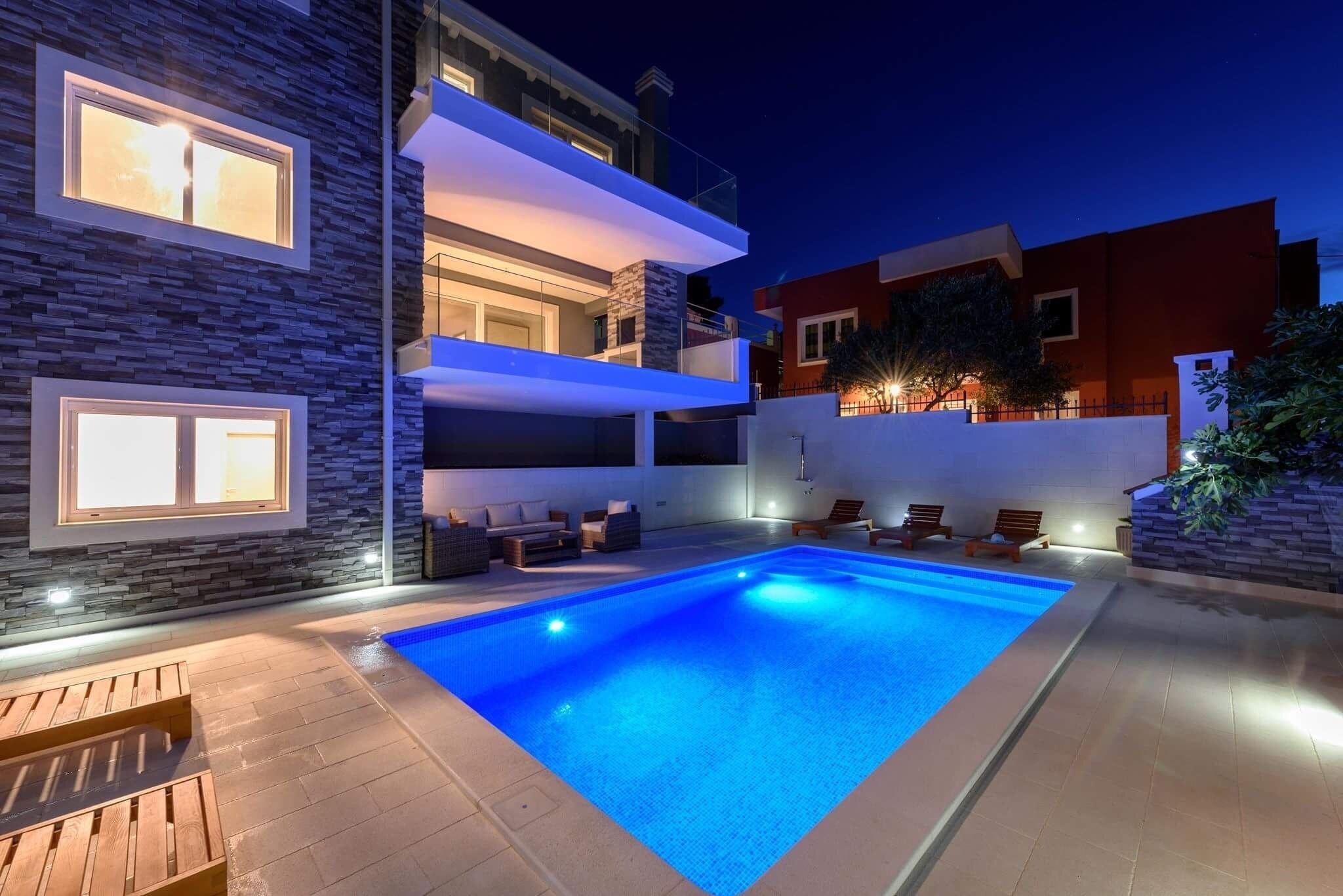 A modern outdoor swimming pool area with a multi-level house in the background.