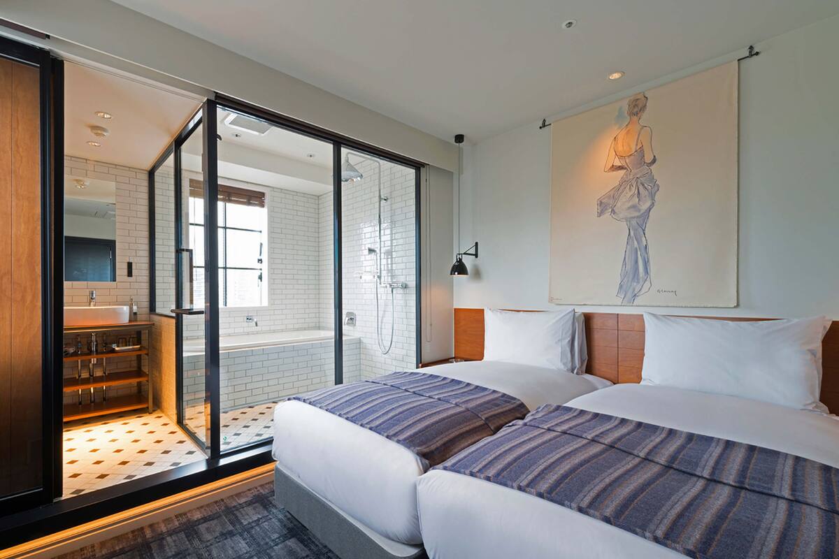 A modern hotel room with a large sliding glass door leading to a bathroom, a bed with striped bedding, and a painting on the wall.