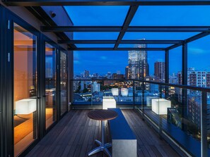 Un balcón moderno con vista a la ciudad, barandillas de vidrio y piso de madera.