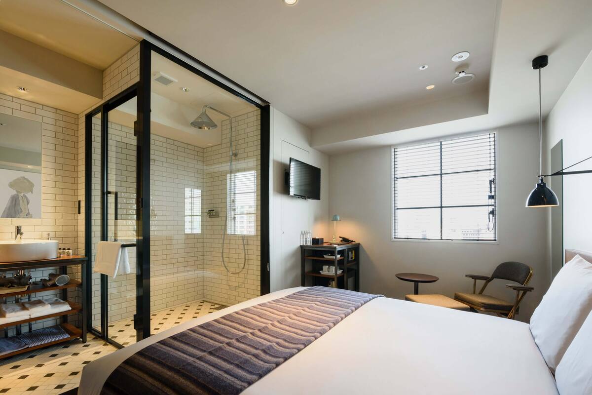 A modern hotel room with a large bed, a glass-enclosed shower, a small desk with a chair, a TV, and a window with blinds.